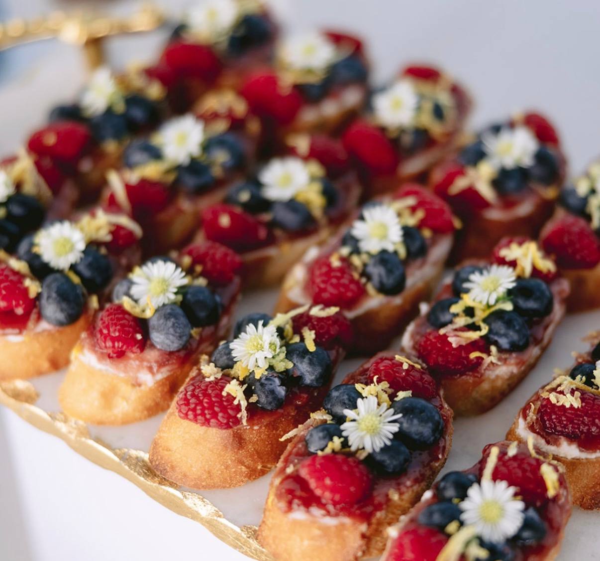 Close-up of berry crostini topped with raspberries, blueberries and edible flowers on a gold tray